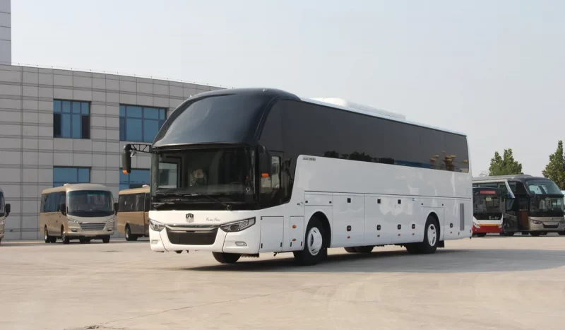 Гладкий белый туристический автобус, напоминающий китайские автобусы Zhongtong LCK6127H в Беларуси, припаркован на бетонной стоянке, на заднем плане вырисовывается современное здание.